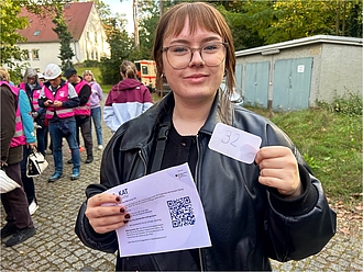 Links im Bild: eine Schlage von Personen; im Vordergrund des Bild eine Person die eine Nummer und einen Infozettel in die Kamera hält